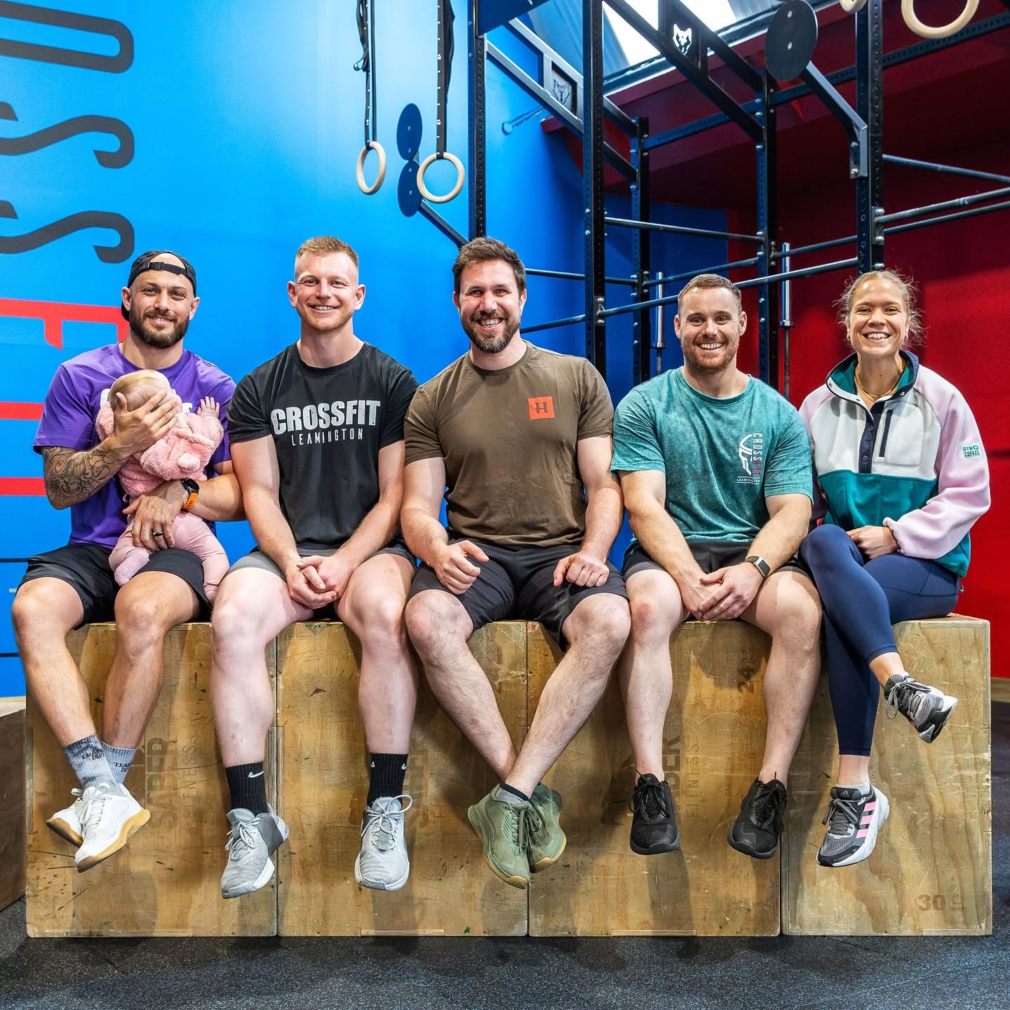 The CrossFit Leamington coaching team laughing together on plyo boxes, one holding a baby