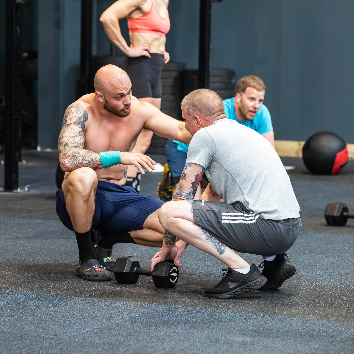 A coach crouching to advise a member holding a dumbbell mid-class